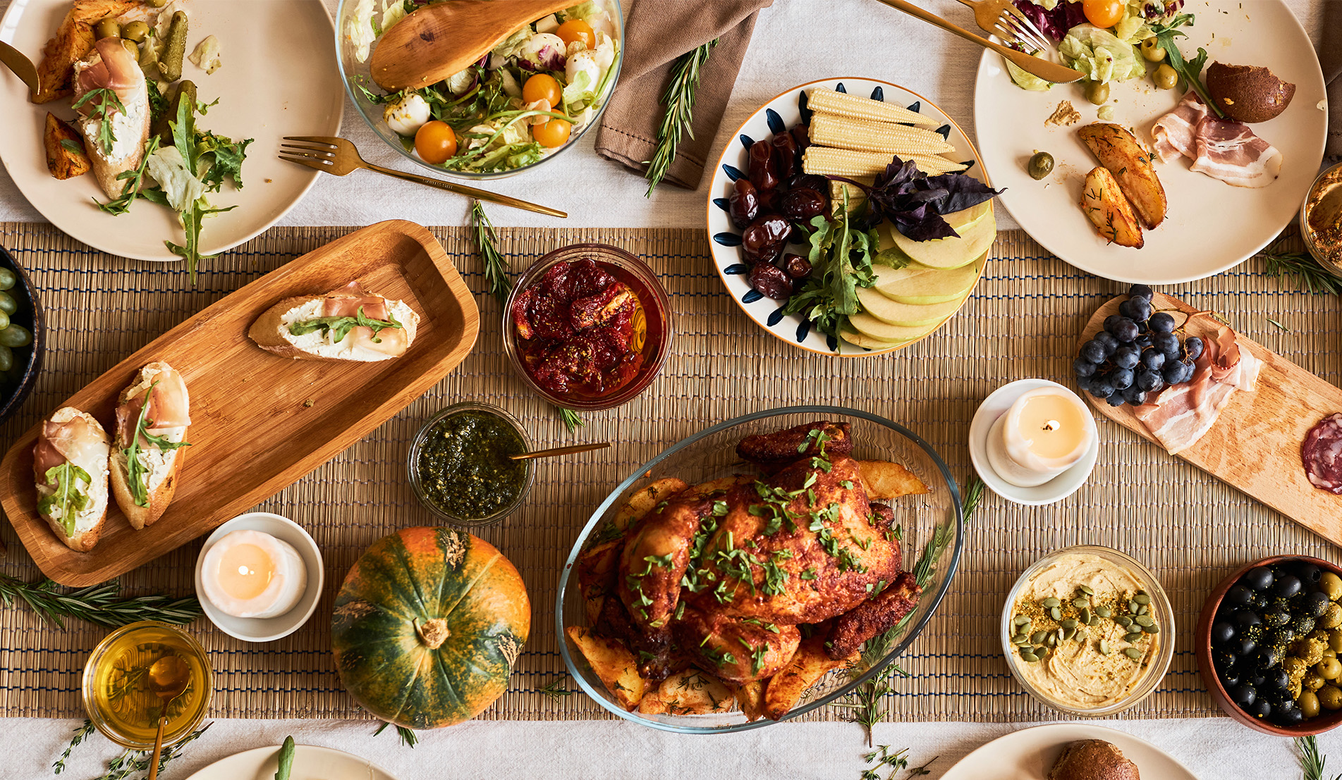 lavish dinner spread including a roast chicken, sundried tomatoes, hummus, olives, charcuterie board, toast points with arugula and prosciutto, and a plate of fresh produce